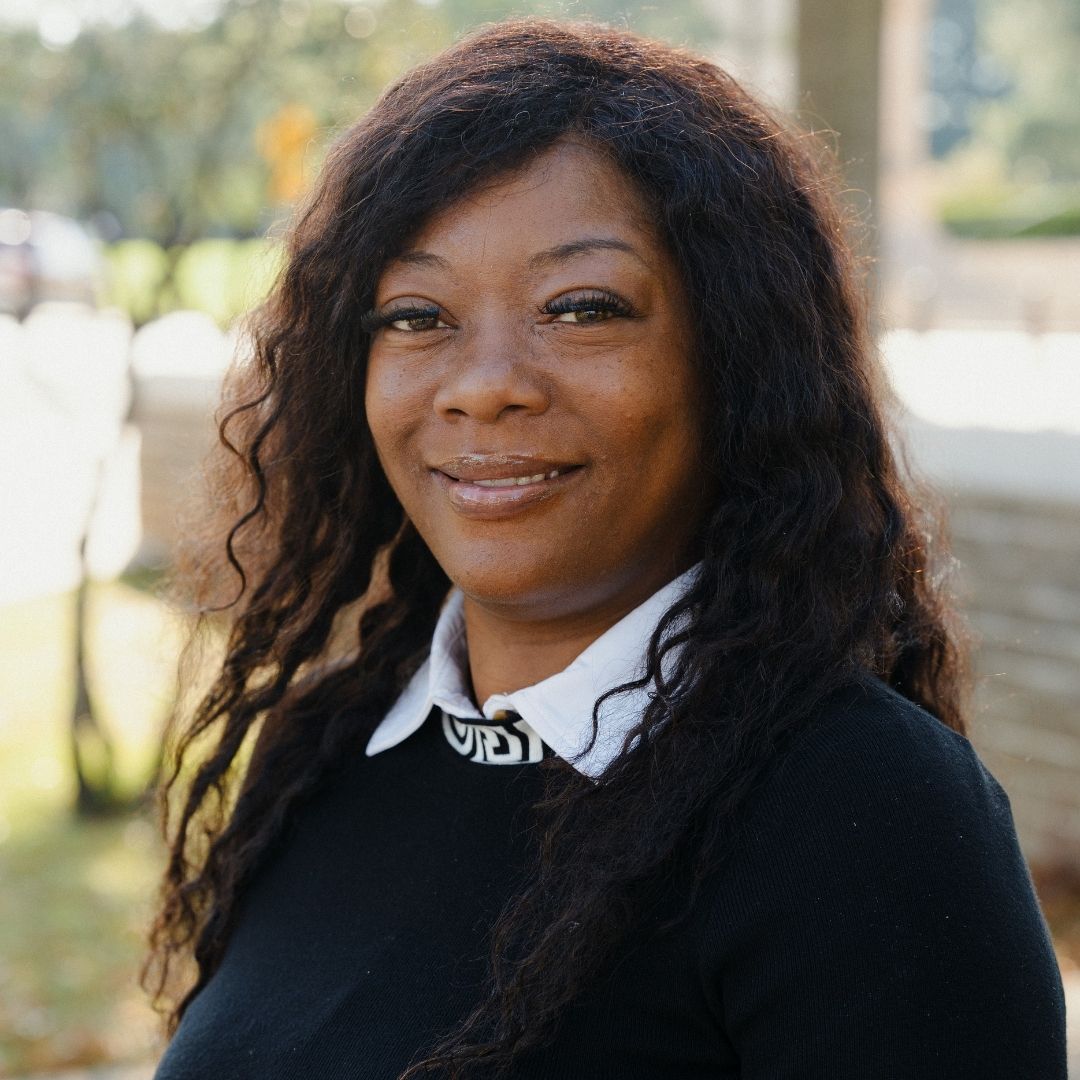 A woman with long hair wearing a black shirt, standing confidently against a neutral background.