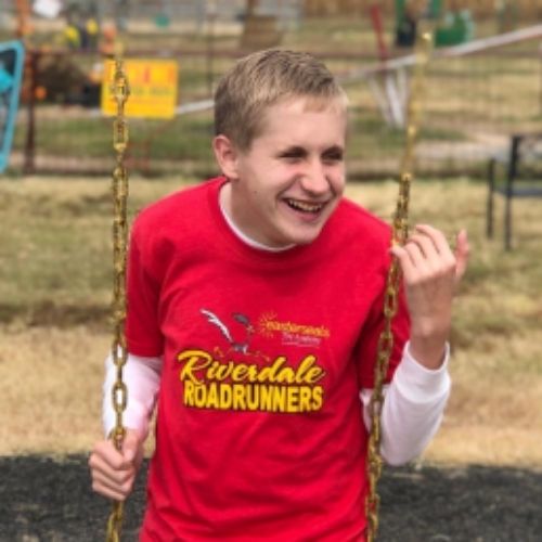 A young boy smiles joyfully while sitting on a swing in a sunny outdoor setting.