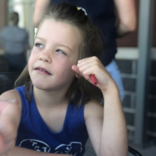 A young girl sits at a table, resting her chin on her hand, deep in thought or contemplation.