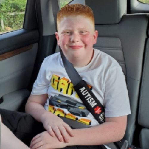 A young boy seated in the back seat of a car, looking at the camera with a thoughtful smiling expression.