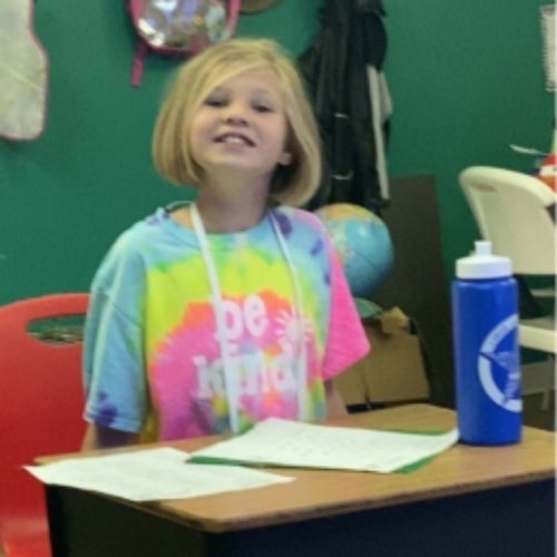 A young girl in a tie dye shirt sits at a desk, focused on her work with a cheerful expression.