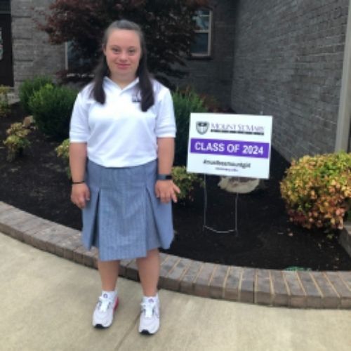 A girl wearing a skirt and white shirt stands in front of a sign, smiling and looking towards the camera.