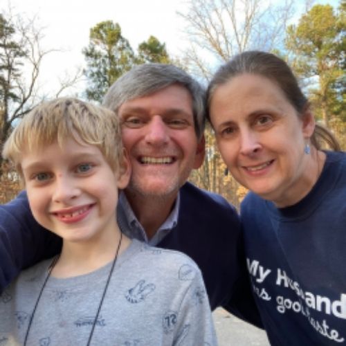 A man and woman stand beside a young boy in front of a large tree, smiling and enjoying a sunny day outdoors.