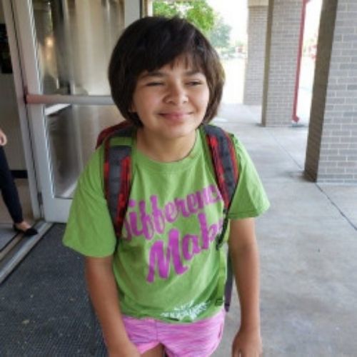 A young girl in a green shirt and pink shorts smiles brightly, enjoying a sunny day outdoors.