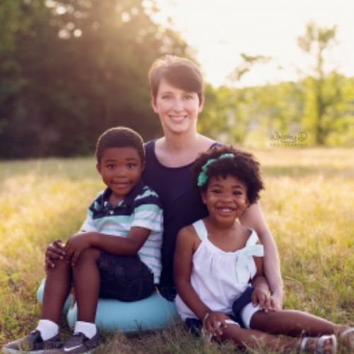 A woman and two children are seated on the grass, enjoying a sunny day together in a serene outdoor setting.