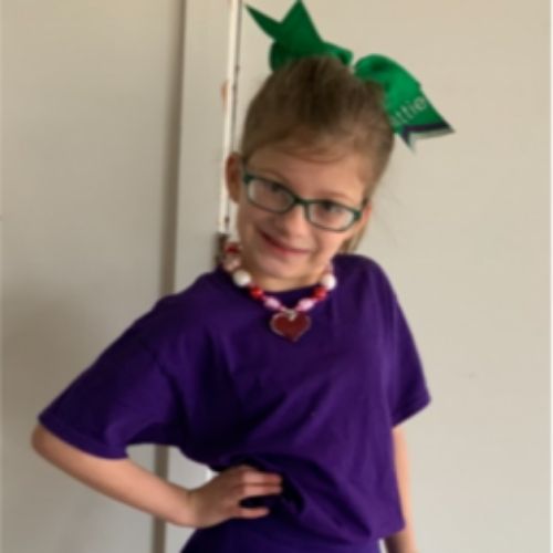 A young girl with glasses smiles while wearing a purple shirt, showcasing her cheerful demeanor and vibrant style.