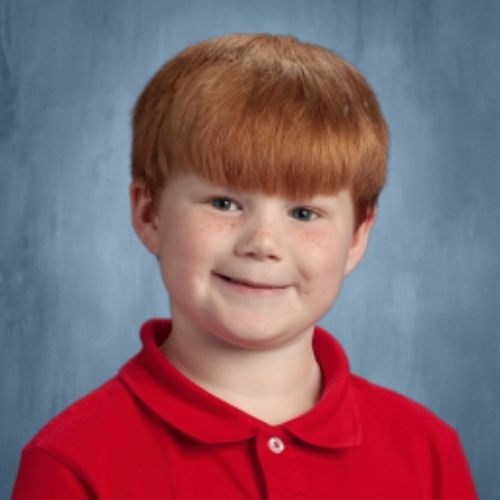 A young boy with vibrant red hair is wearing a bright red shirt, smiling cheerfully in a sunny outdoor setting.