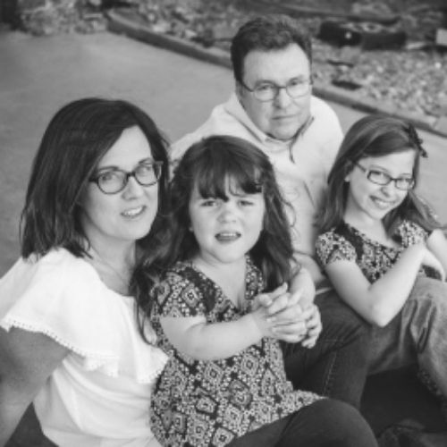 A family sits together on the ground, enjoying a moment with their daughters, surrounded by nature and warmth.