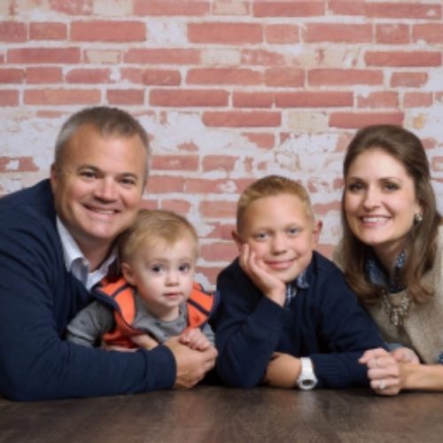 A family stands together smiling for a photo in front of a textured brick wall.