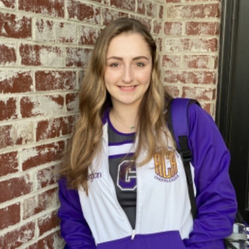 A young woman wearing a stylish purple and white jacket, standing confidently with a pleasant expression.