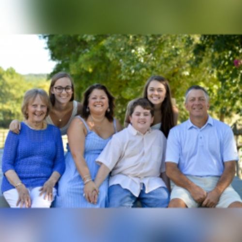 A family stands together wearing varying shades of blue and white, smiling for a photo in front of a large, leafy tree.