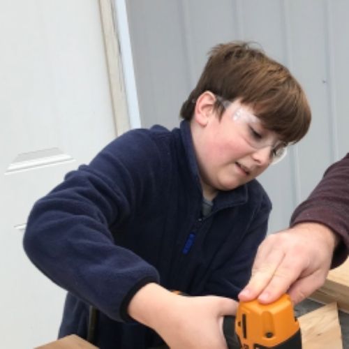 A young boy operates a drill, focused on constructing a wooden box with precision and care.