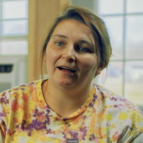 A woman in a tie-dye shirt sits, surrounded by warm colors and homey decor.