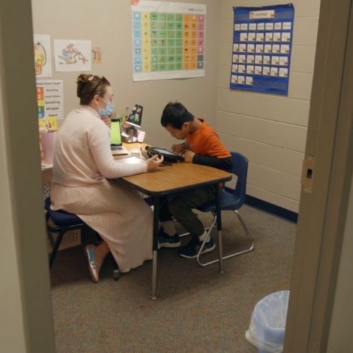 A woman and a child are seated at a desk in a classroom, engaged in a learning activity together.