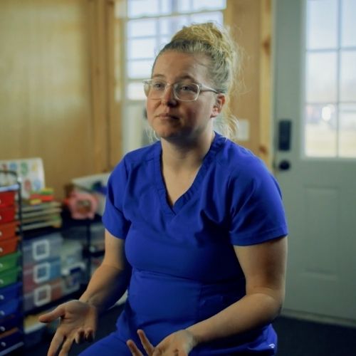 A woman wearing blue scrubs is seated in a room, appearing focused and engaged in her surroundings.