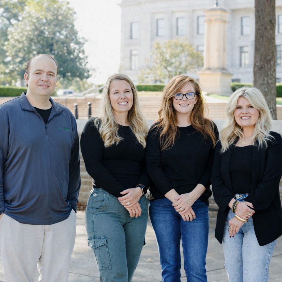 Four adults—one man and three women—stand outdoors in front of a historic-looking building. They are smiling and dressed in casual clothing, with trees and steps visible in the background.