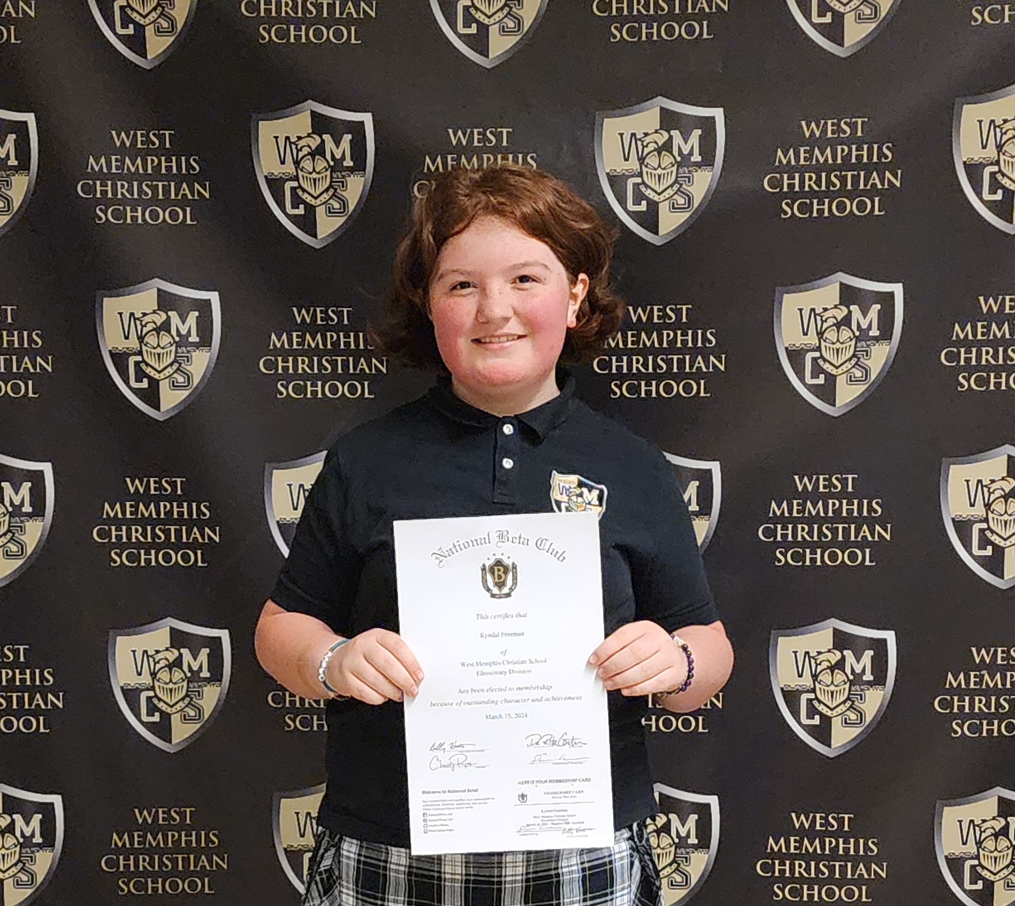 A smiling student in a black polo shirt and plaid skirt holds a National Beta Club certificate in front of a backdrop with the West Memphis Christian School logo.