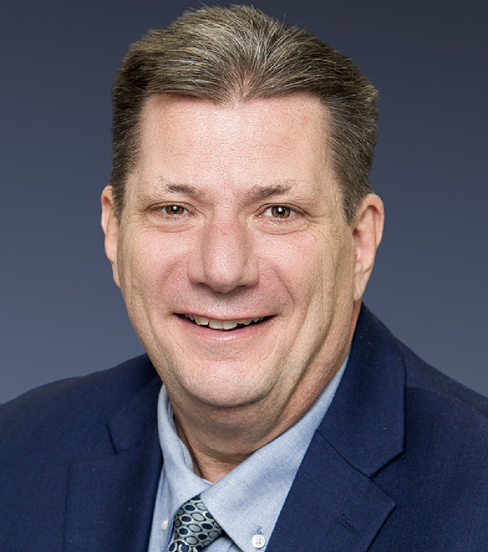 A middle-aged man with short brown hair, wearing a navy blue suit jacket, light blue shirt, and patterned tie, smiles at the camera against a plain dark background.