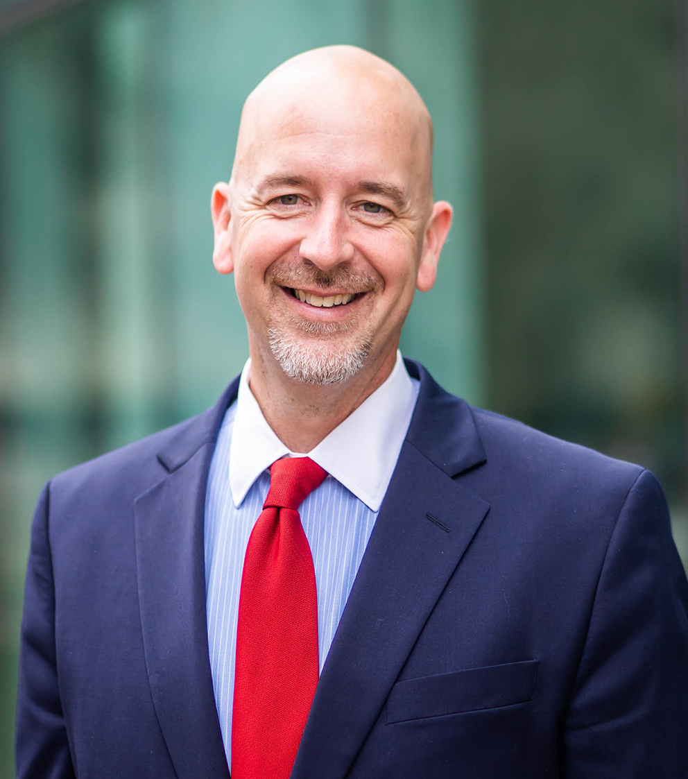 A bald man with a goatee is smiling while wearing a navy blue suit, blue striped shirt, and red tie, standing in front of a blurred green and gray background.
