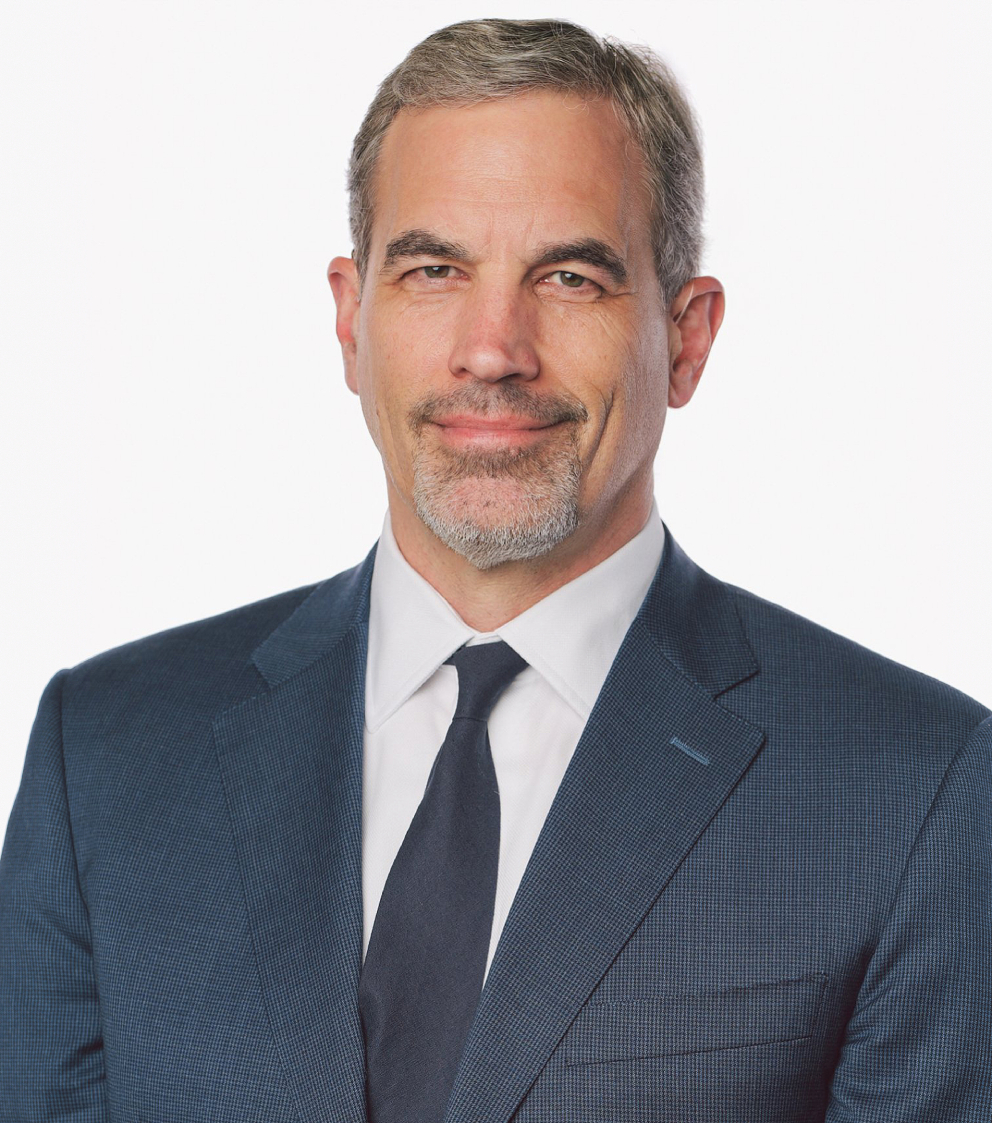 A middle-aged man with short gray hair and a goatee, wearing a blue suit, white shirt, and dark tie, smiling slightly against a plain white background.