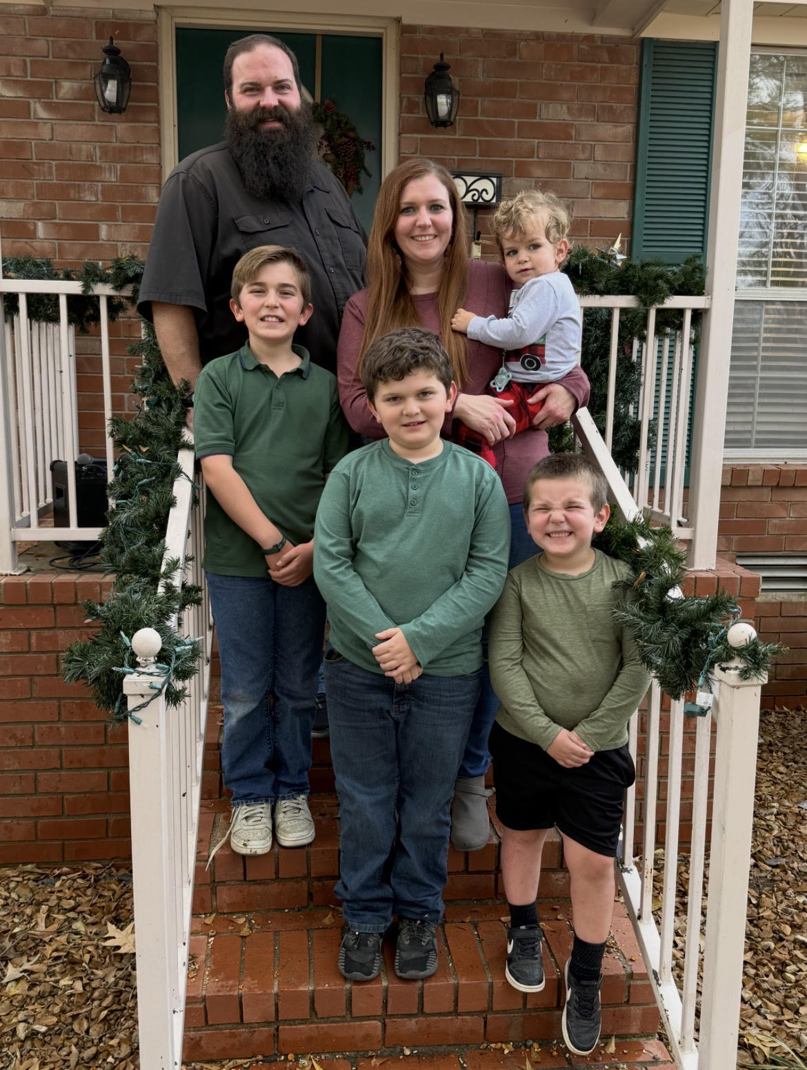 Screenshot A family of six poses on brick steps decorated with green garland. Two adults stand behind four children, one holding a toddler. The group smiles in front of a house with a brick wall, white railing, and green shutters.