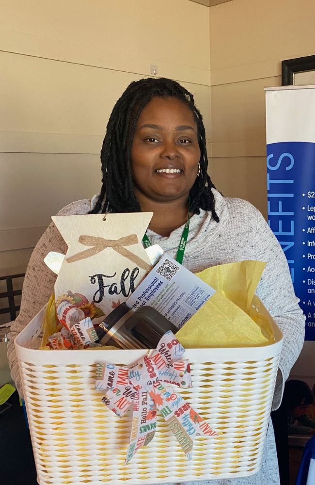 A woman with long braids smiles while holding a white gift basket filled with goodies, including a Fall sign, ribbon, and yellow tissue paper. A blue and white sign is partially visible in the background.