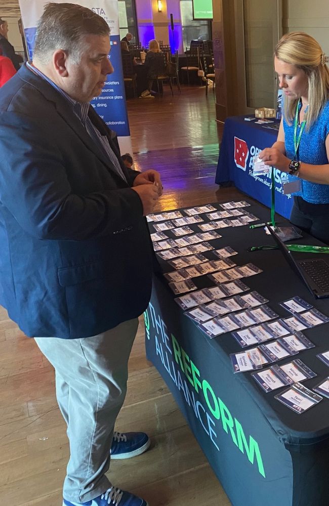 A man in a blue blazer stands at a table covered with name tags, speaking to a woman behind the table at an event. The tablecloth reads THE REFORM ALLIANCE and other booths are visible in the background.