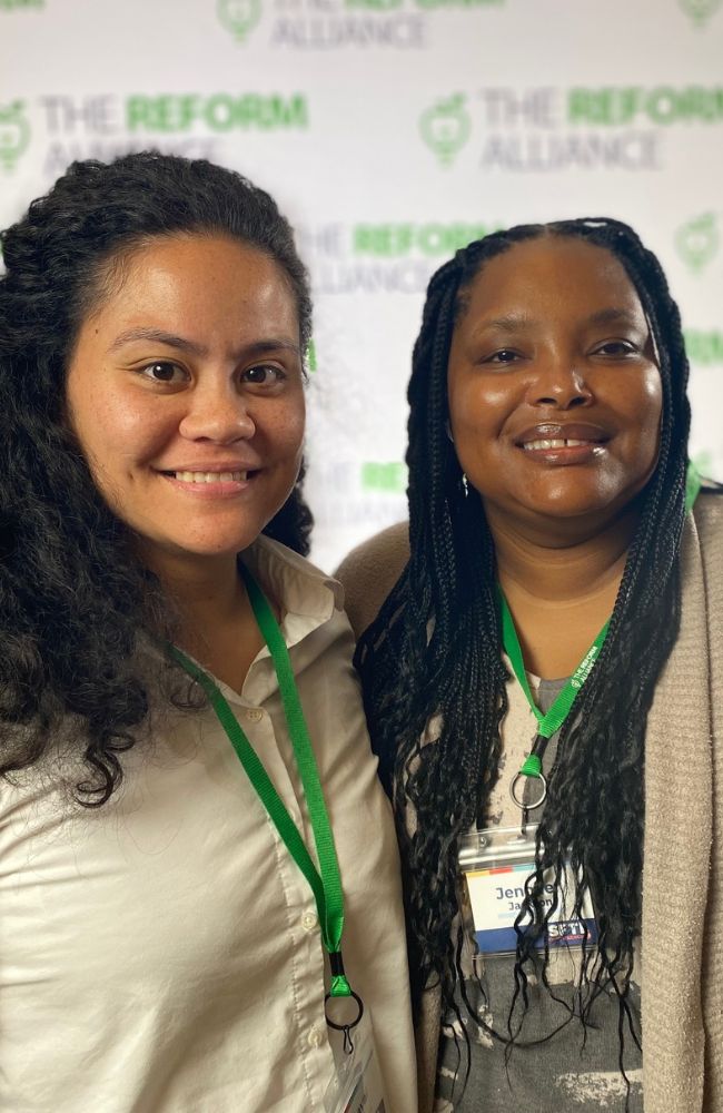 Two women wearing green lanyards and smiling stand in front of a backdrop with The Reform Alliance logo partially visible in the background.