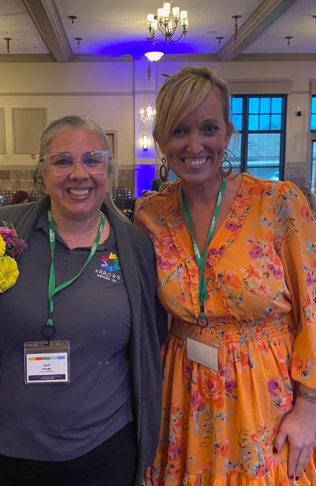 Two women smiling indoors at an event. One wears a gray polo with a name badge and glasses, while the other wears an orange floral dress. Both have green lanyards, and chandeliers hang from the ceiling behind them.