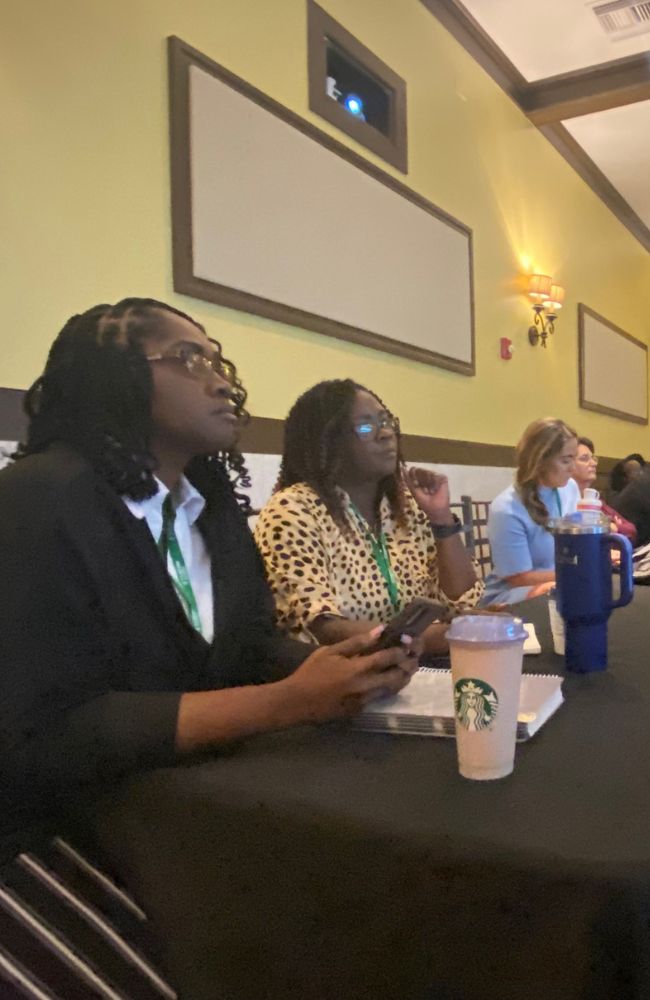 Several people sit at a long table in a conference room, listening attentively. Two women in the foreground have notebooks and drinks, including a Starbucks cup, and appear focused on a presentation out of the frame.