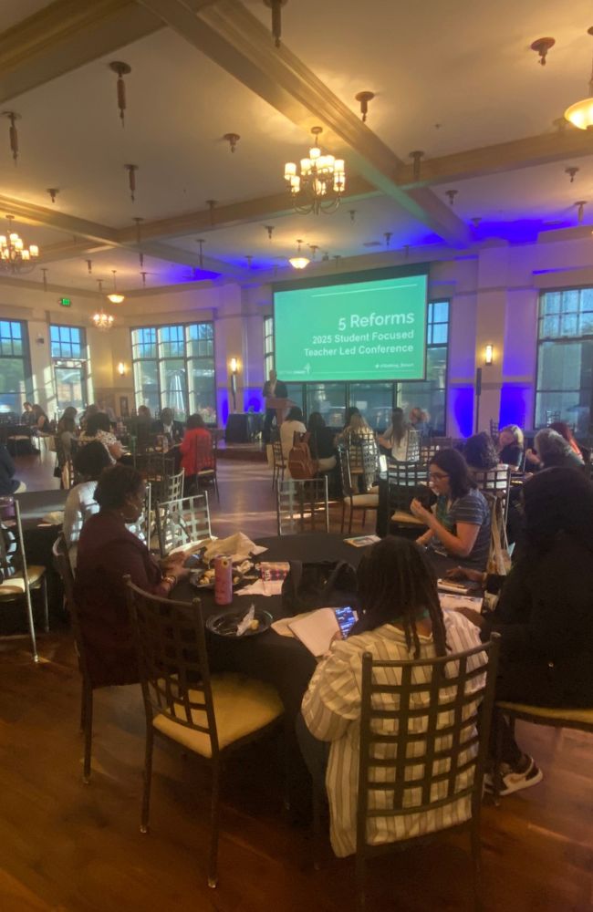 Large room with round tables and people seated, watching a presentation. A screen at the front displays 5 Reforms: 2025 Student Focused Teacher Led Conference. Blue lights illuminate the stage area.