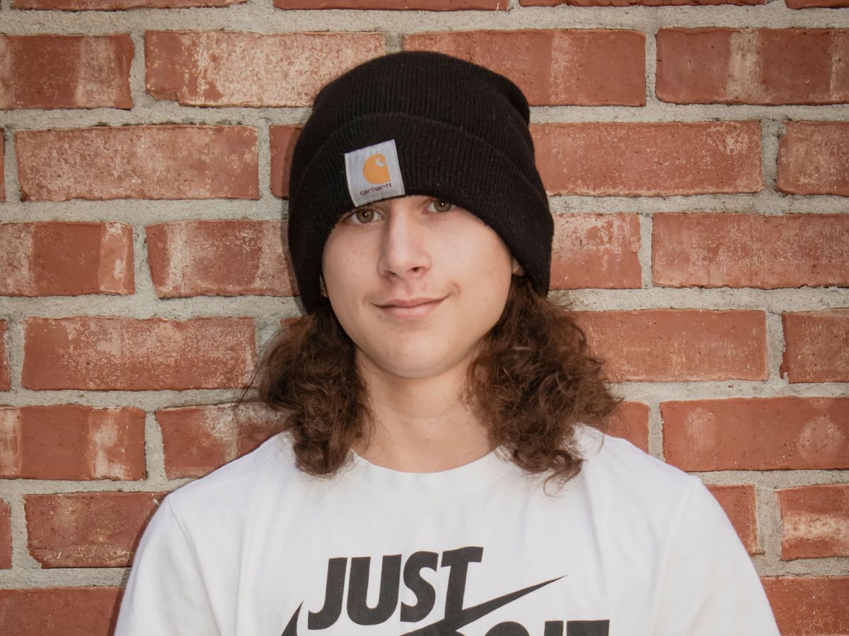 Faces of Freedom – The Boucher Family A person with long, curly brown hair wears a black beanie and a white Just Do It t-shirt, standing in front of a red brick wall.