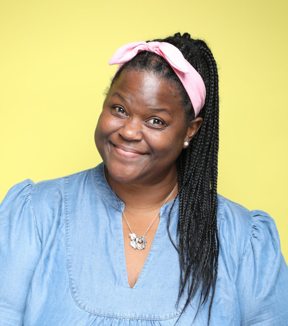 Smiling woman with long braided hair wearing a pink headband, blue blouse, and necklace, standing in front of a yellow background.