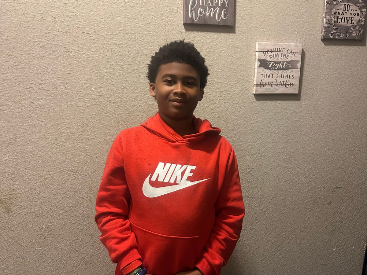 Untitled design.jpgFaces of Freedom – The McIntosh Family A boy wearing a red Nike hoodie stands in front of a beige wall decorated with framed quotes and signs. He is smiling softly and has his hands clasped in front of him.