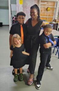 Four people stand together indoors, smiling. An adult woman in a black dress and three children in black outfits, two boys with colored ties and one blonde girl holding her dress, pose happily for the photo.