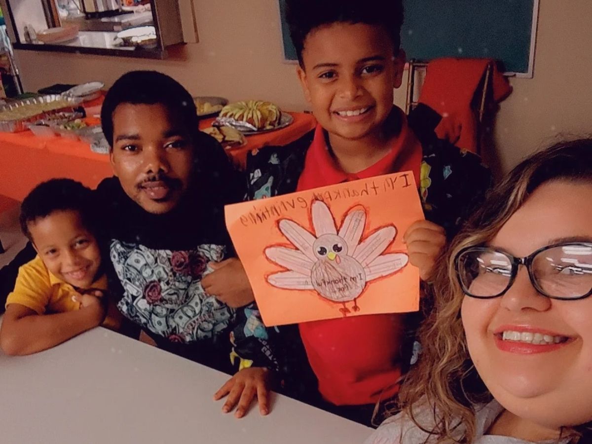 Faces of Freedom – The Coffman Family Four people smile at a table; a child in red holds a hand-drawn Thanksgiving turkey. Food is set out on a table in the background, and everyone appears happy and cheerful.