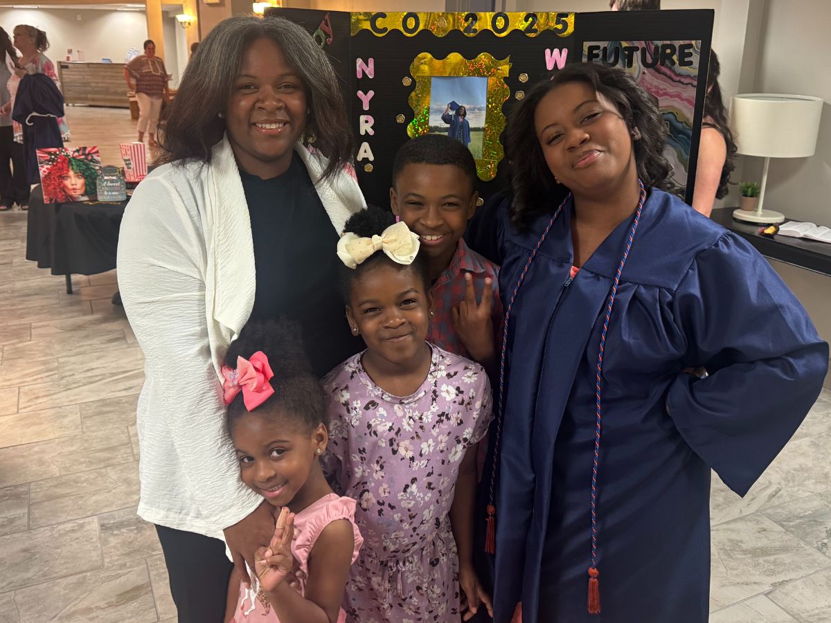 Faces of Freedom – The Taylor Family A woman and four children smile at a graduation event; one child wears a blue cap and gown. A decorated board with photos and the words CONGRATS NYRA 2023 is displayed in the background.