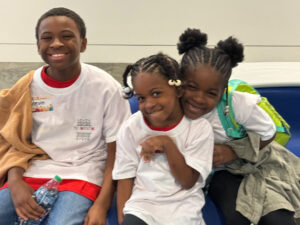 Three smiling children sit close together on a blue bench. They wear white shirts, and one child holds a water bottle. Two girls have braided hair, and the boy has short hair and a tan blanket on his shoulder.