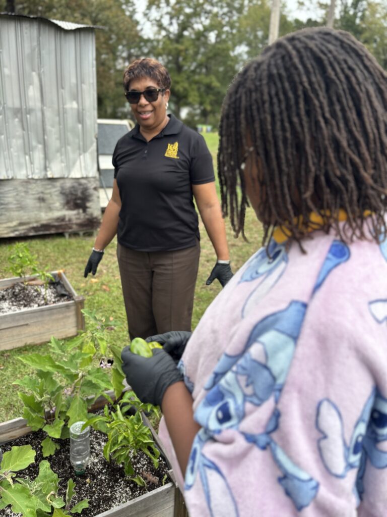 A woman and a girl wearing black gloves pick vegetables from a raised garden bed.